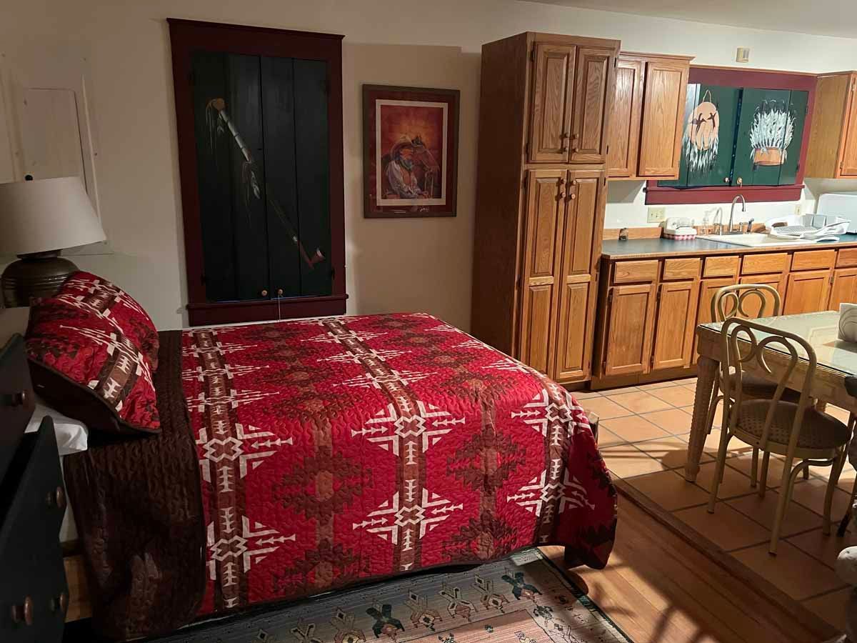 Bedroom with bed, red patterned quilt, open kitchen with wooden cabinets.