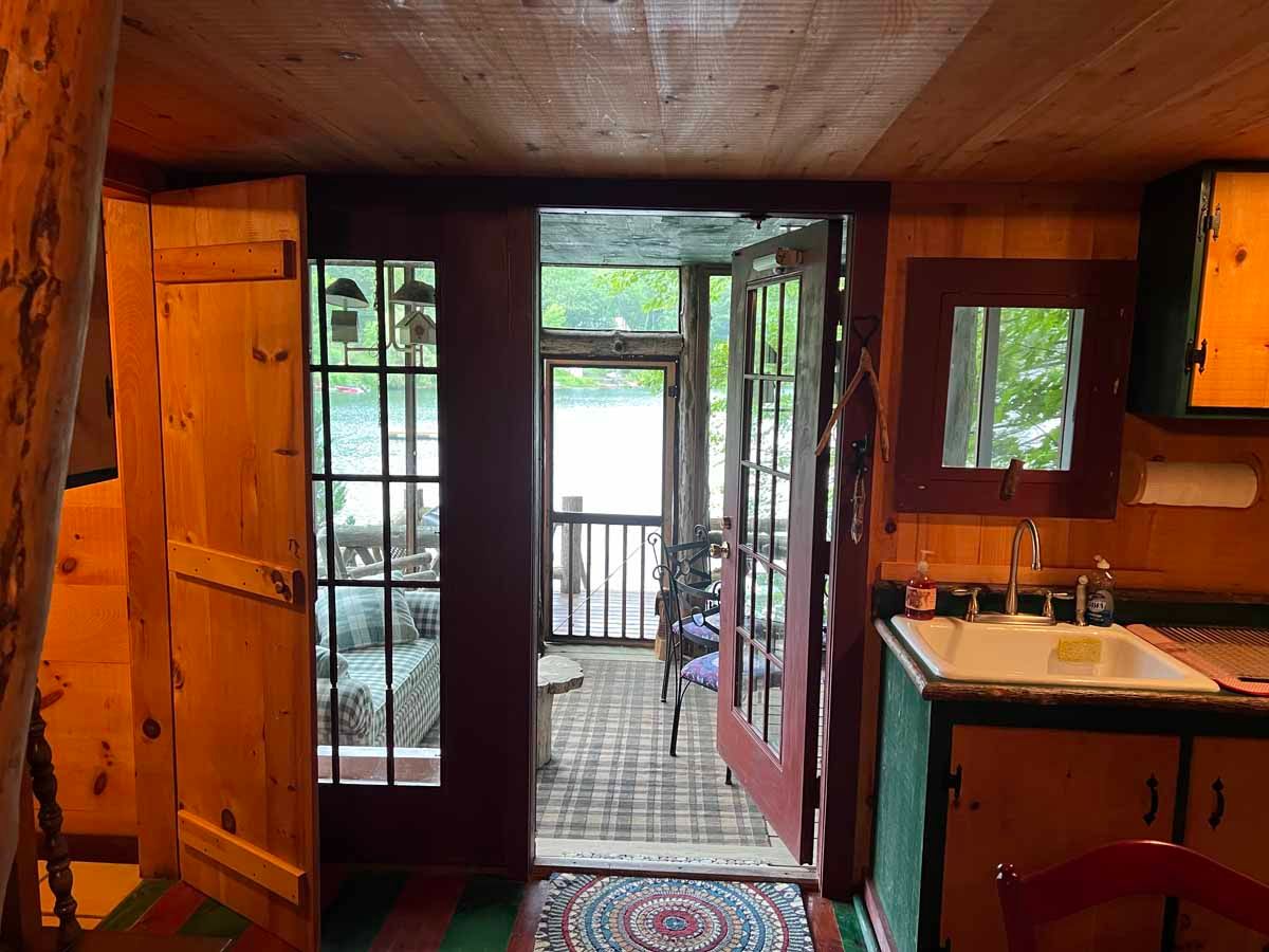 Interior of a cabin with open doors leading to a porch overlooking water; rustic wood and colorful details.