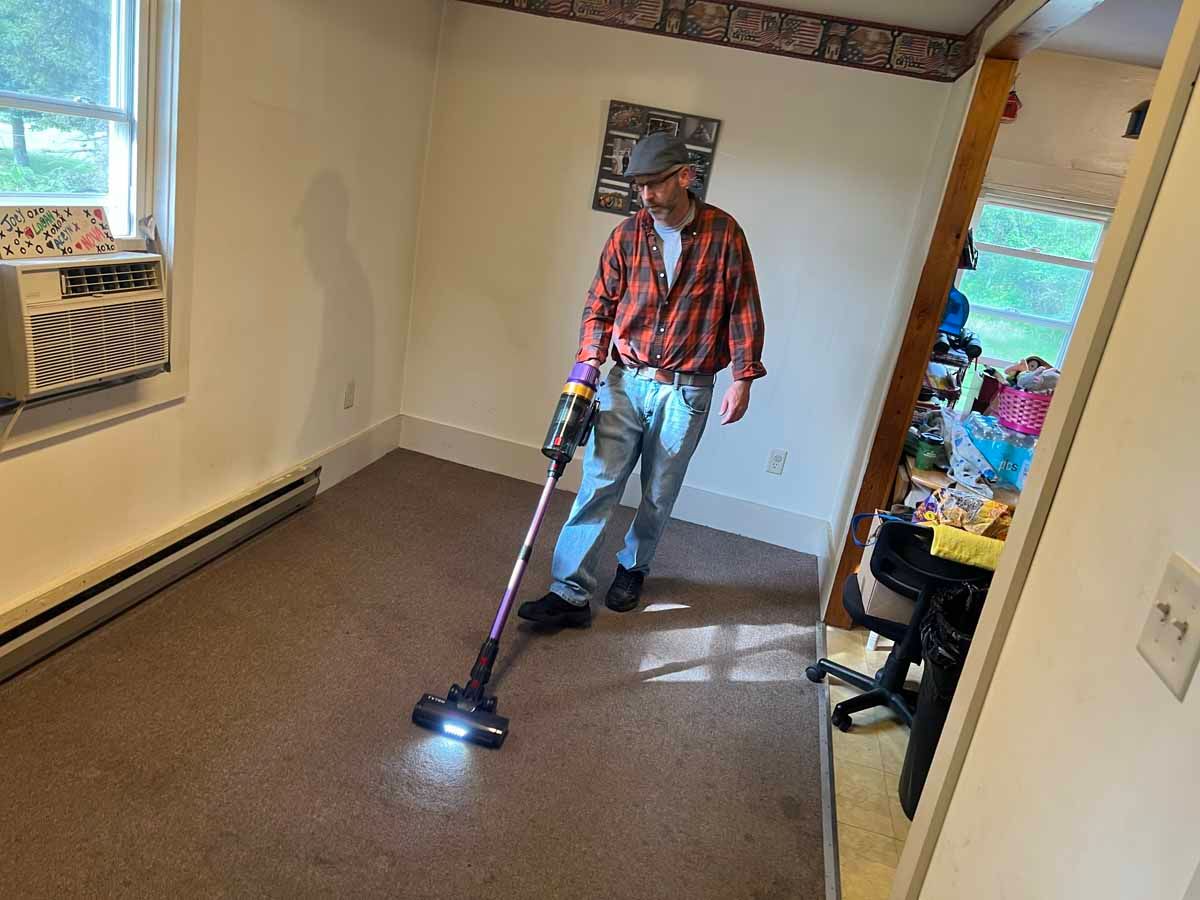 Man vacuums a carpeted room; he's wearing a plaid shirt and jeans. A window and doorway are visible.