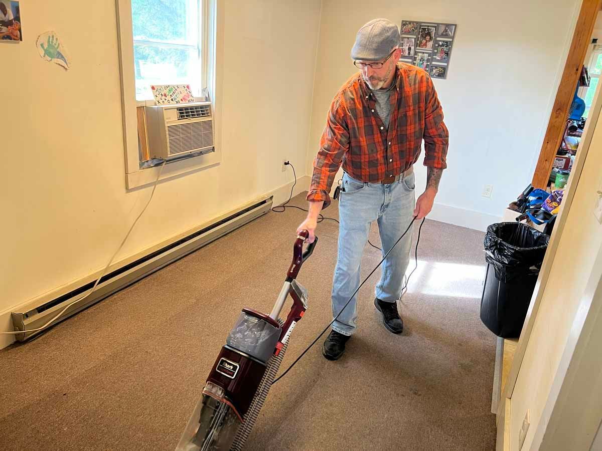 Man vacuums a carpeted room. He wears a plaid shirt, jeans, and a cap. The room has a window and baseboard heaters.