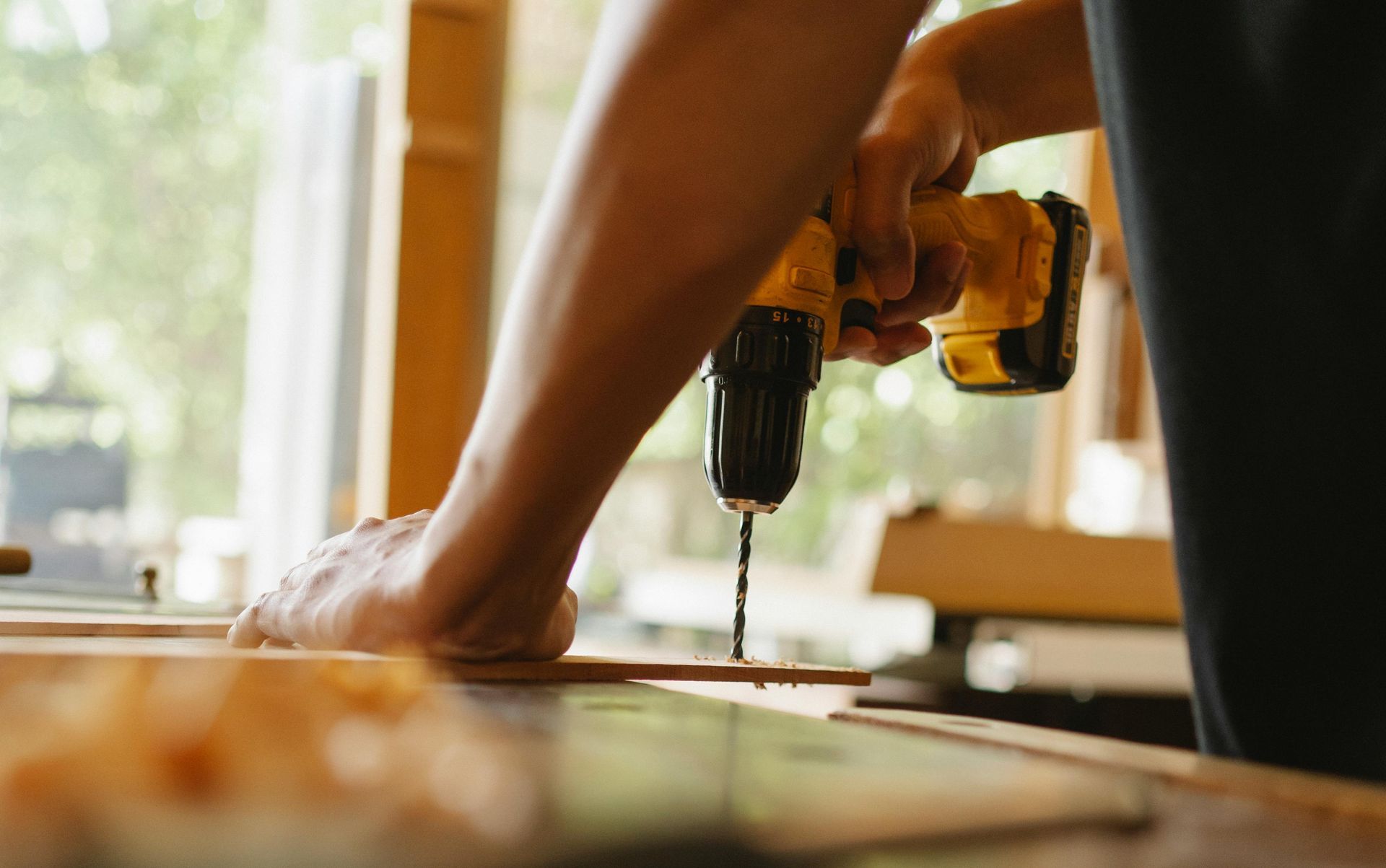 Person using a yellow cordless drill to drill into a wooden board in a workshop.