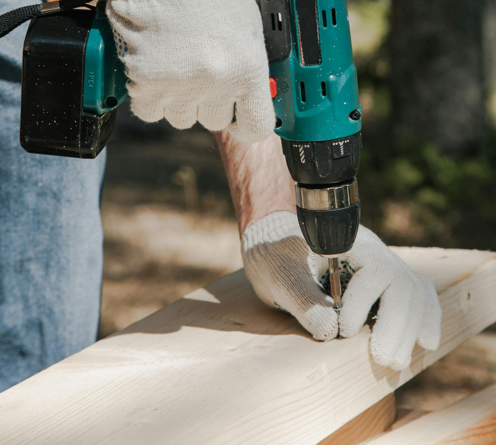 Person using a power drill on a wooden floor, wearing a green work glove, near a small, square hardware piece.