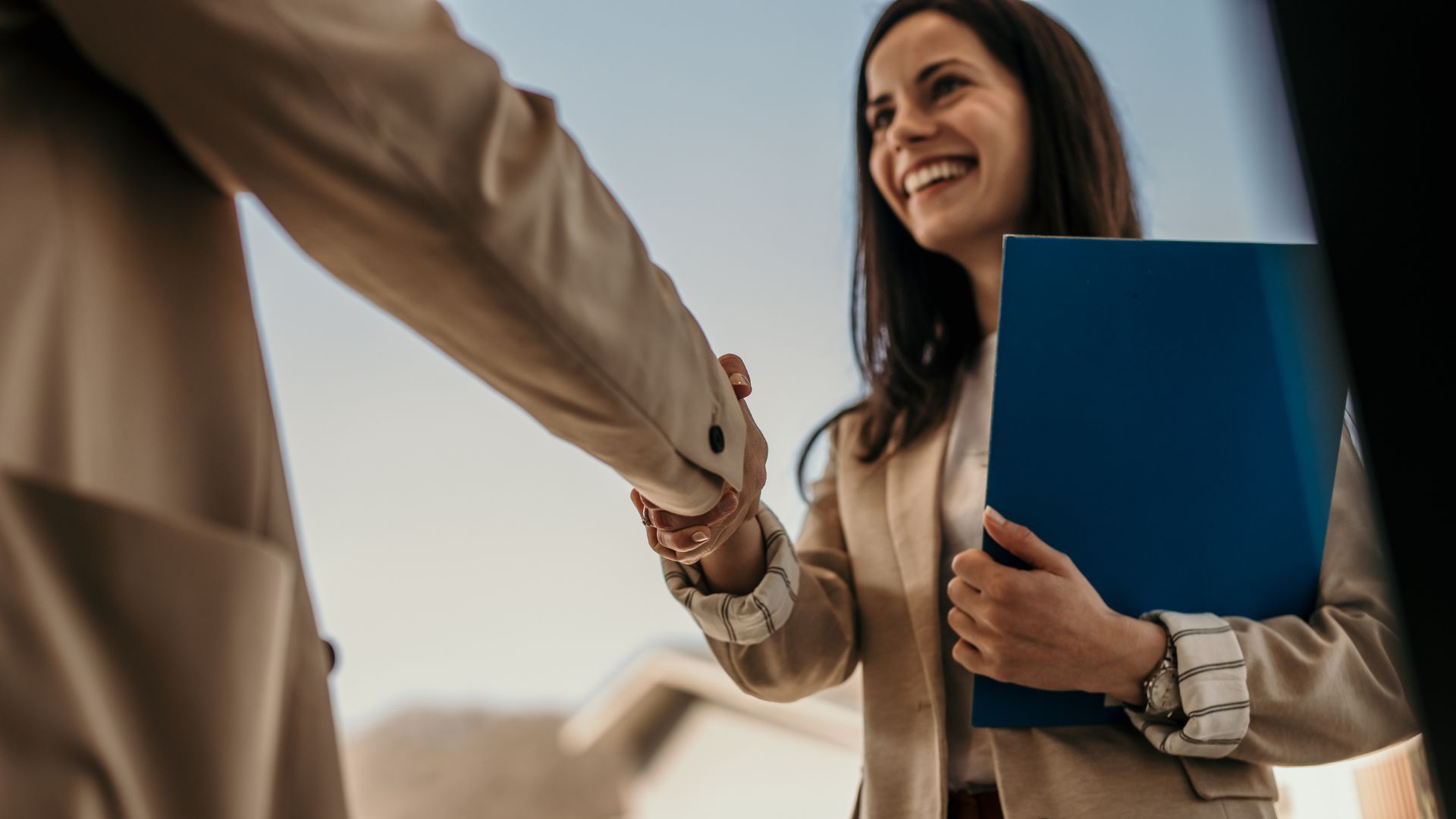 Woman in beige blazer smiles, shaking hands with another person. She holds a blue folder, outdoors.