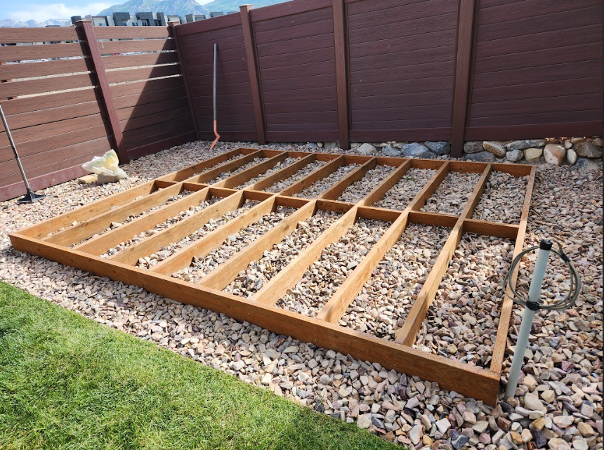 A wooden rectangular frame for a deck or shed foundation sits on a bed of gravel in a backyard next to a dark wood fence.