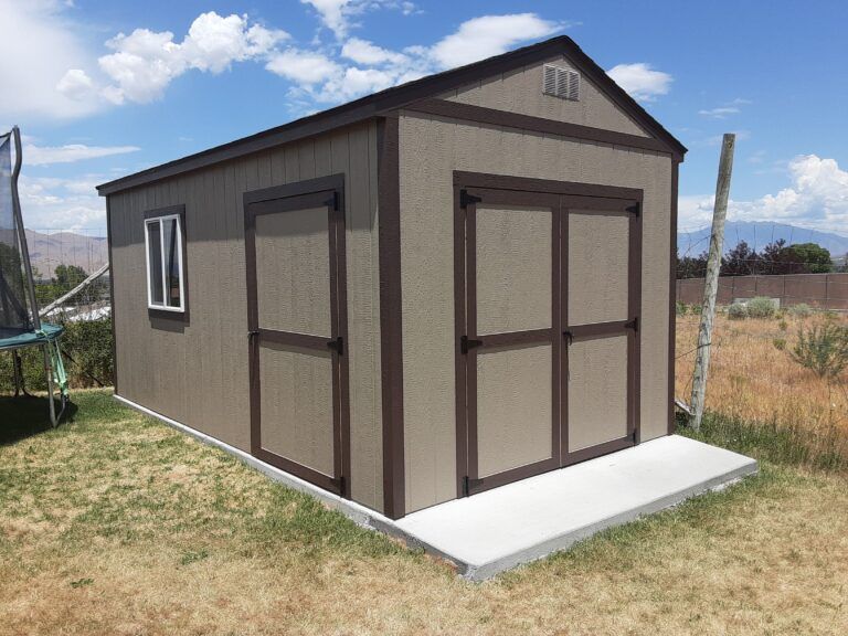 Tan and brown shed with double doors and window on a concrete slab, set in a grassy yard under a blue sky.