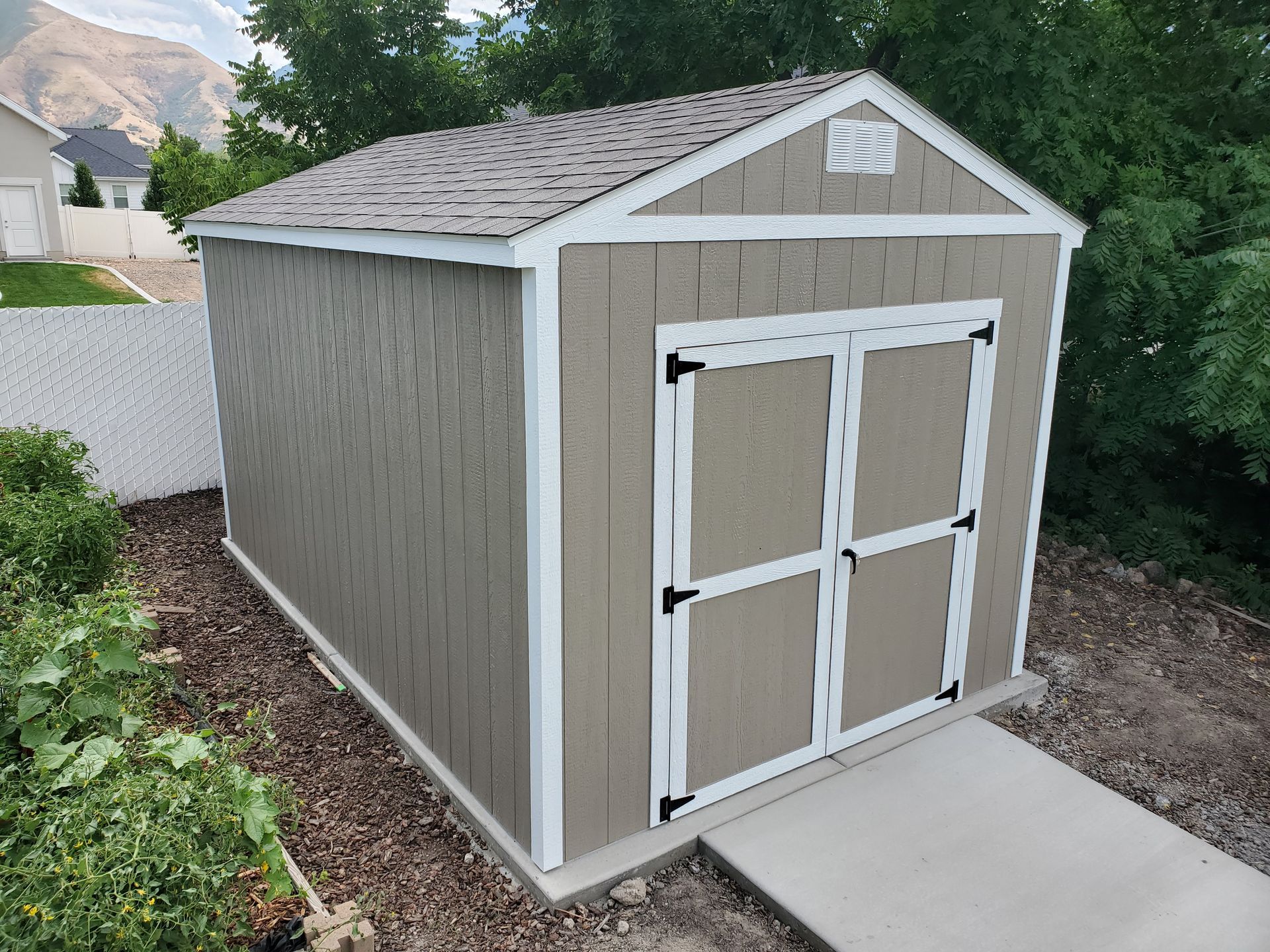A tan storage shed with white trim and grey shingled roof sitting on a concrete pad in a yard.