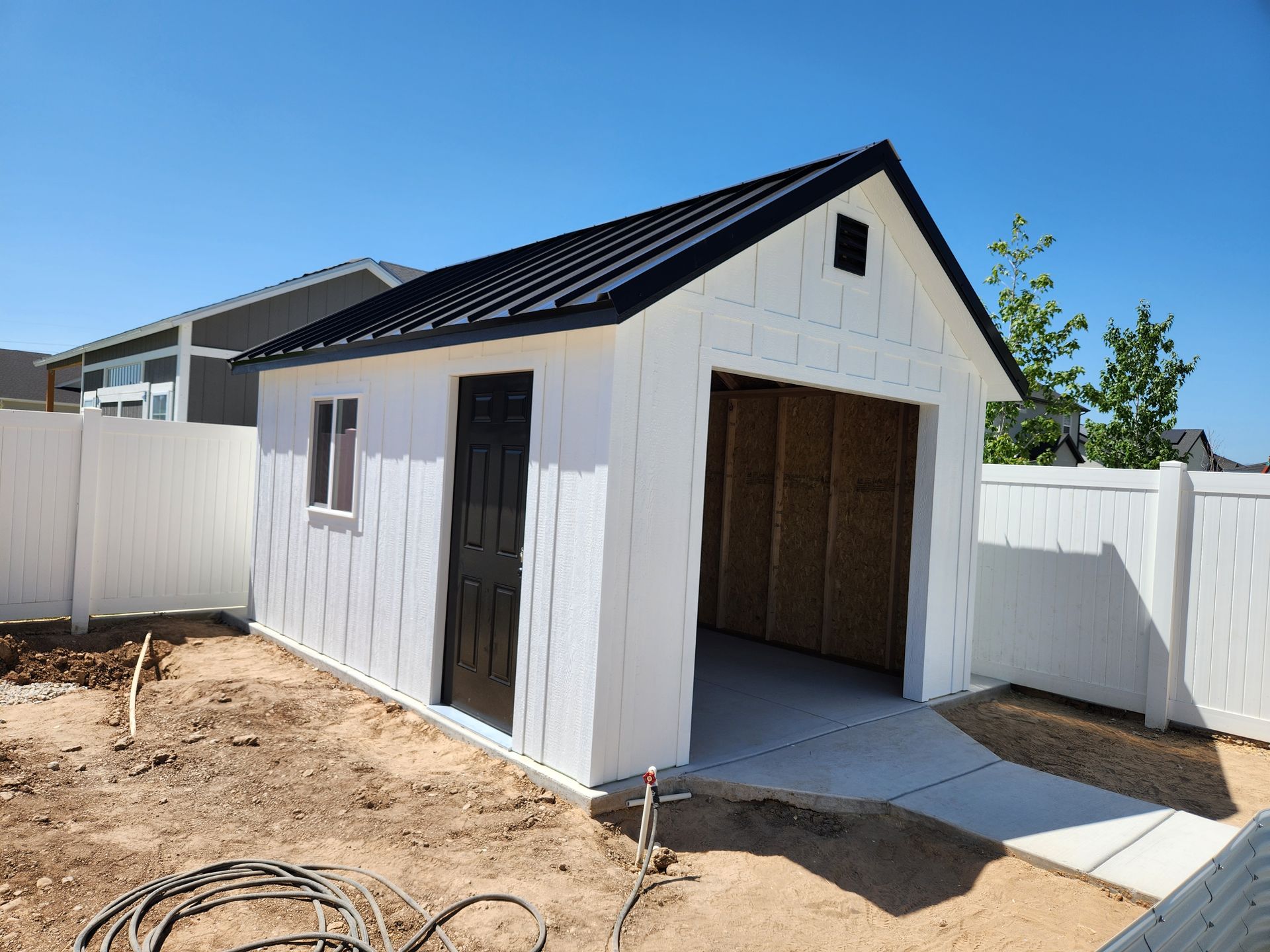 A new white shed with a black metal roof and door stands in a backyard with white vinyl fencing and dirt ground.