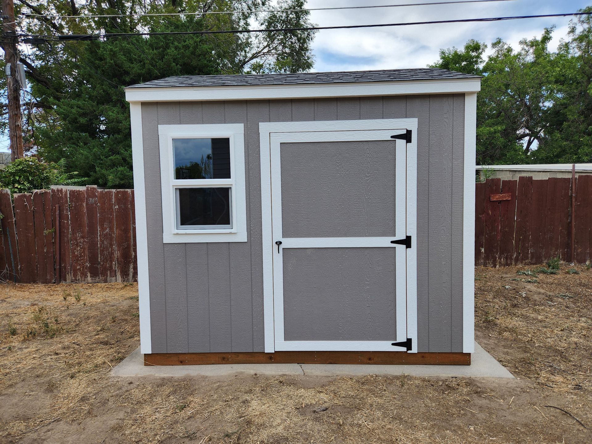 A grey wooden storage shed with white trim, a single window, and a door, sitting on a concrete pad in a fenced yard.