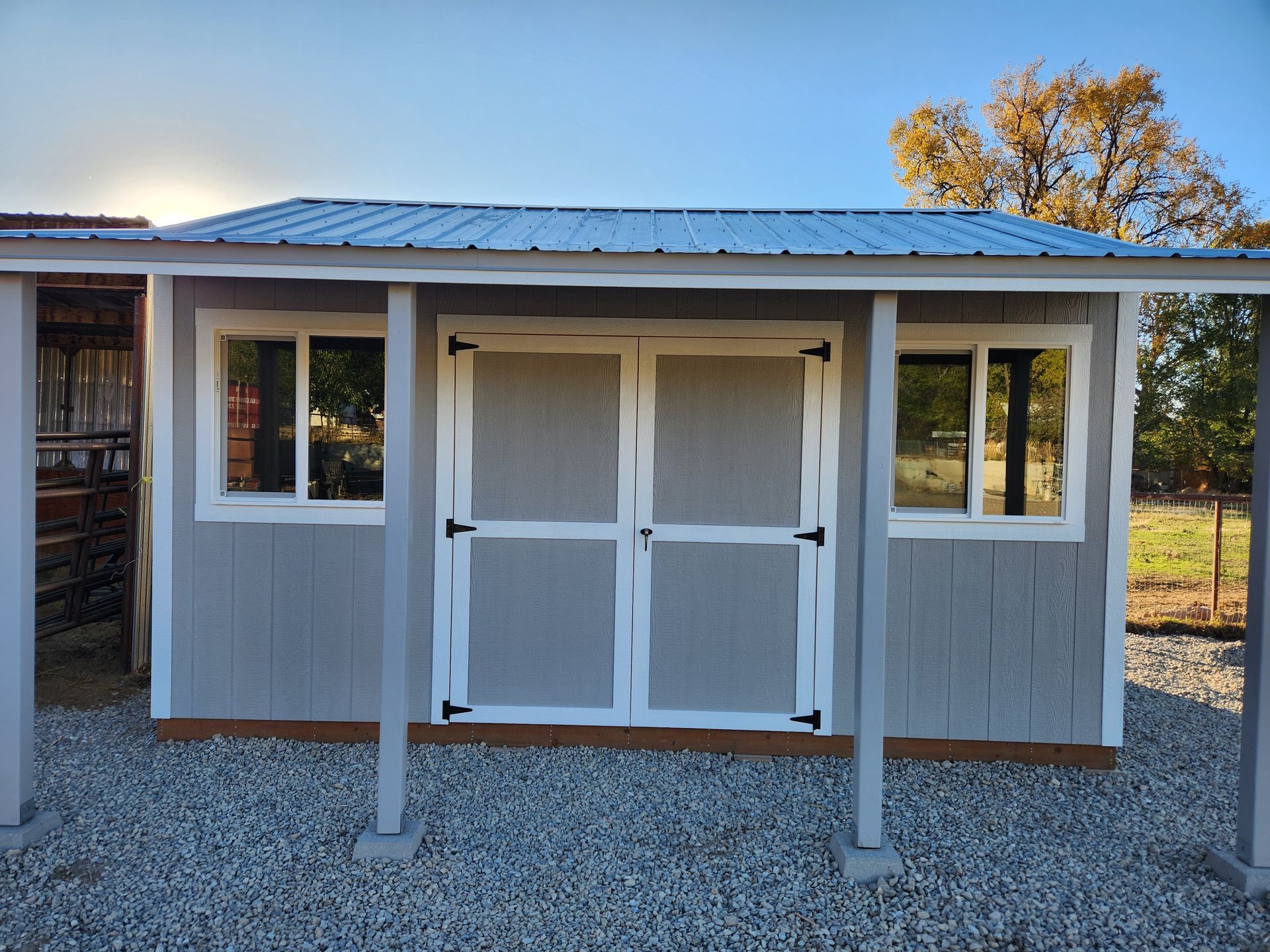 A grey, rectangular shed with white trim, a metal roof, and double doors, situated on a gravel surface outdoors.