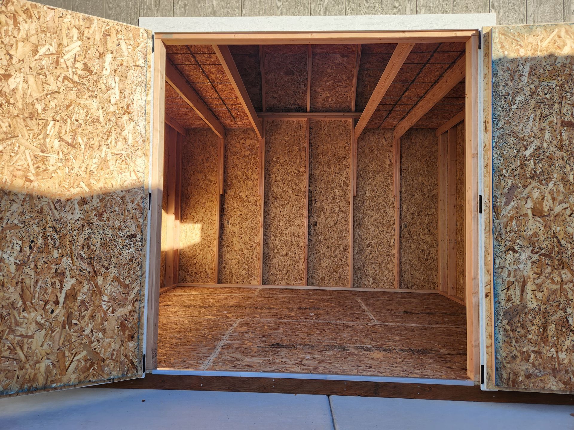 The open, wide-angle doorway of an empty, unfinished wooden shed with plywood walls and floor.