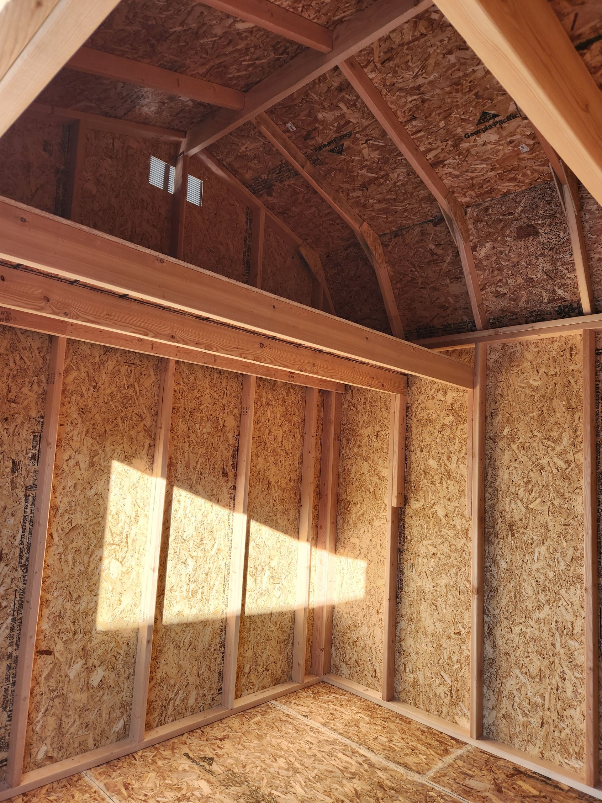 Interior view of an unfinished wood shed with a loft, showing framed walls and rafters with natural light.