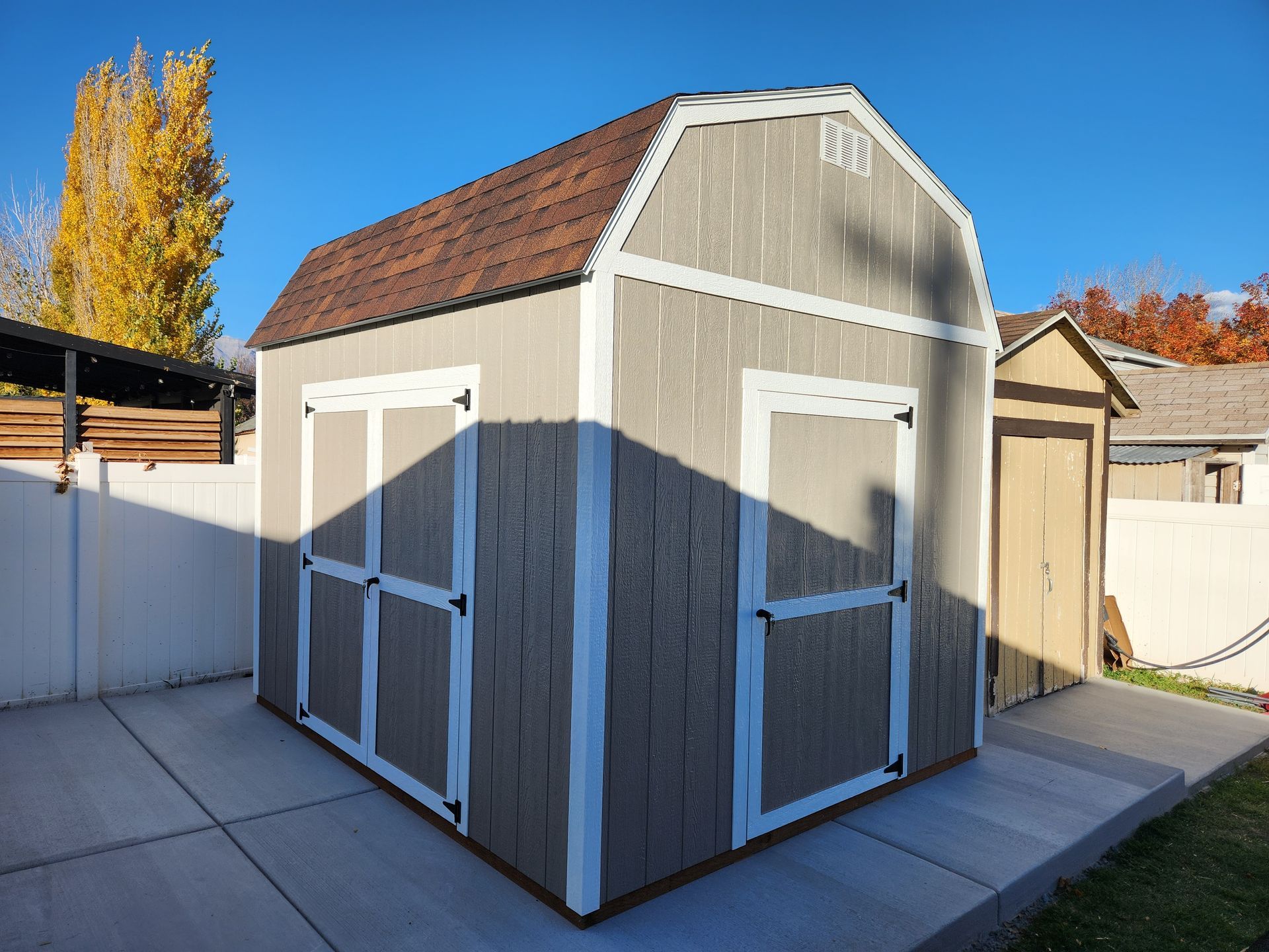 A tan gambrel-roof shed with white trim and double doors sitting on a concrete patio in a backyard on a sunny day.