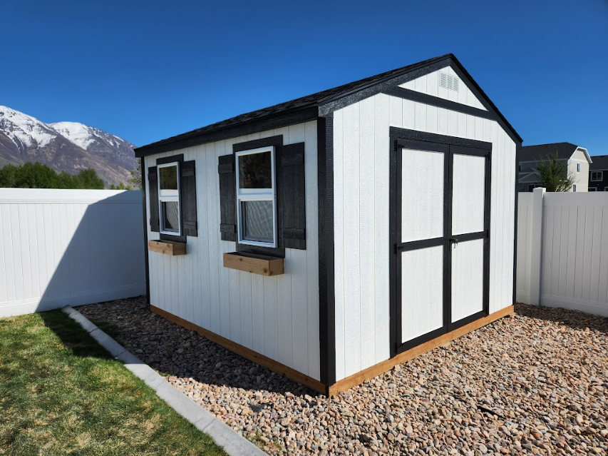 A white backyard shed with black trim and shutters, two windows with flower boxes, and double doors on a gravel base.