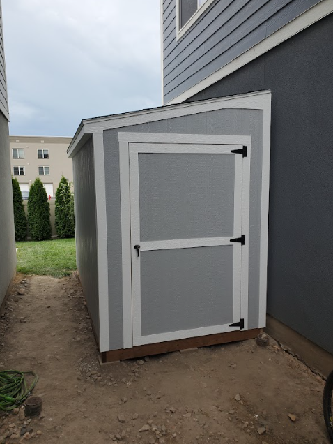 A gray, single-door lean-to shed with white trim positioned in a narrow outdoor side yard between two houses.