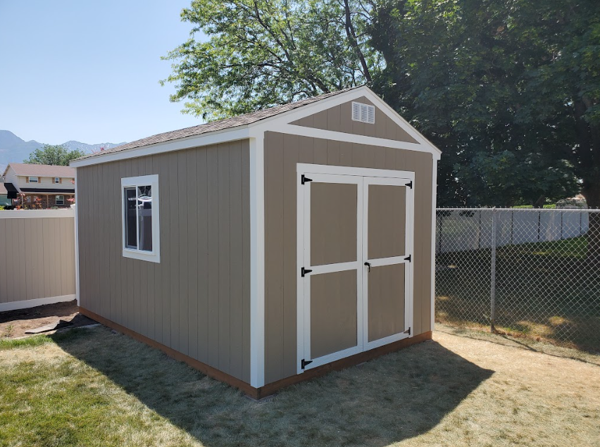 A tan storage shed with white trim, double doors, and a window, situated in a backyard with fences and a mountain view.