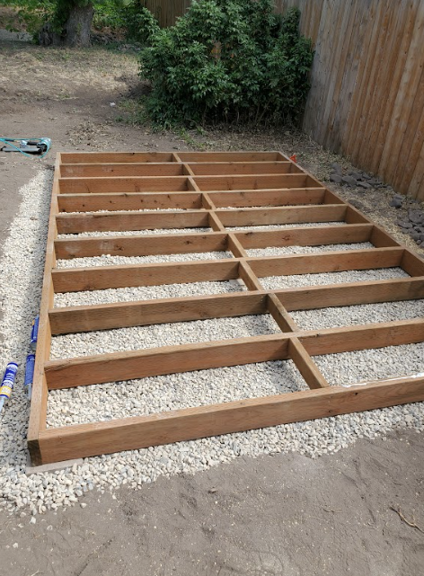 A wooden rectangular deck frame sits on a gravel-filled foundation next to a wooden fence.