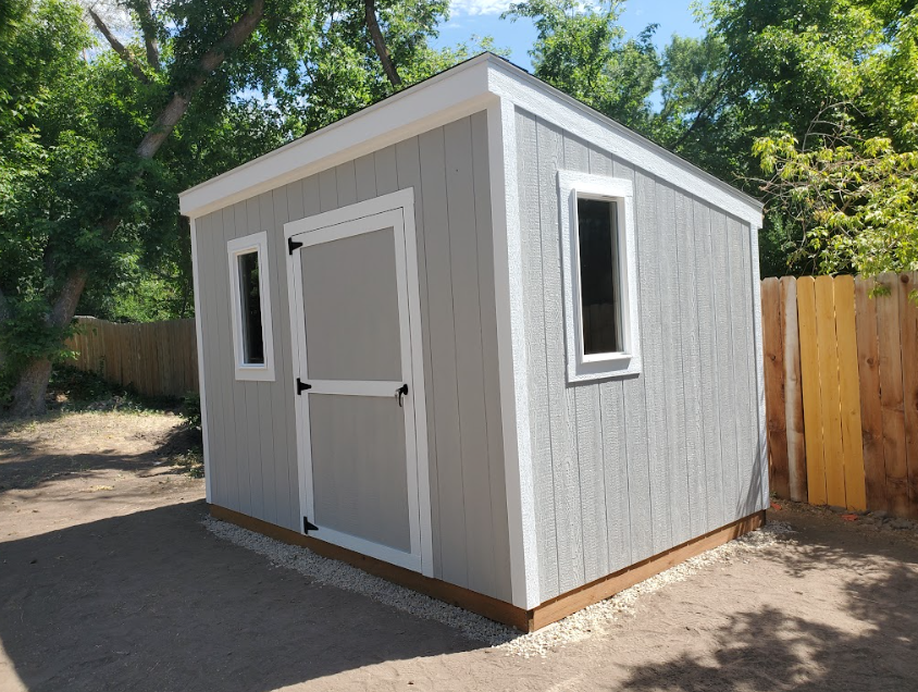 A light gray backyard shed with white trim, a single door, and two windows, set in a sunny, wooded outdoor area.
