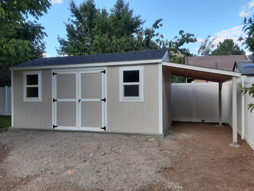Tan shed with white trim, double doors, two windows, and an attached open-sided wooden carport in a residential yard.