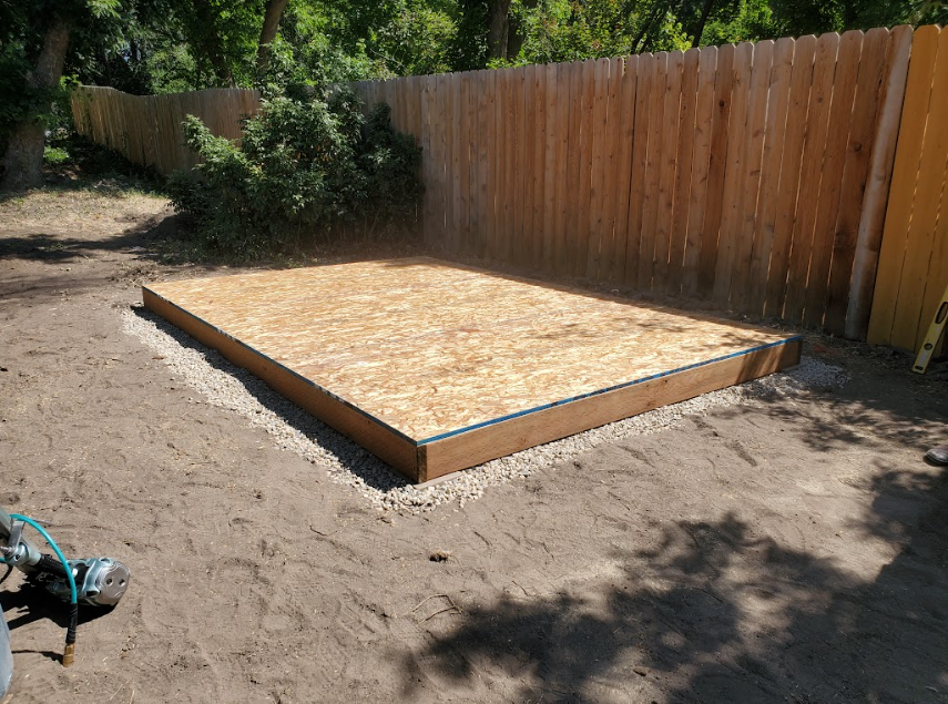 A rectangular wooden shed foundation base sits on a bed of gravel in a yard next to a tall wooden fence.
