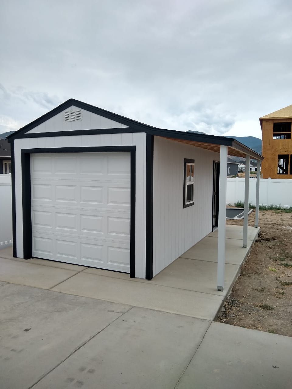 A small, white residential shed with a black trim, a garage door, a side window, and a small covered porch.