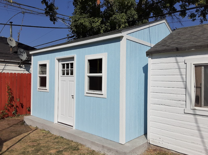 A light blue shed with a white door and two windows sits on a concrete foundation in a backyard.