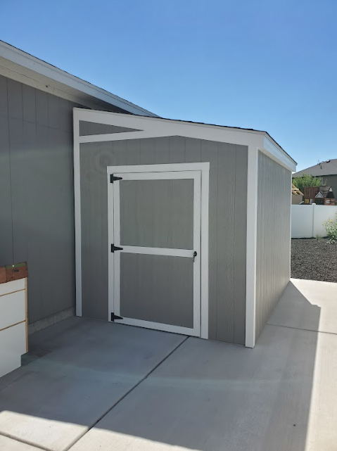 A gray storage shed with white trim and a single door attached to the side of a house on a concrete patio.