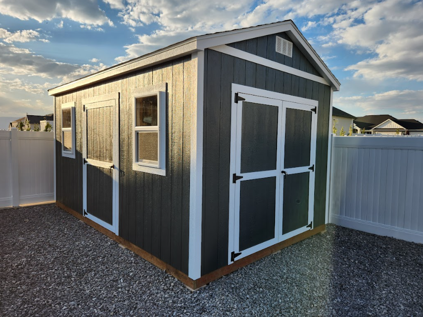 A dark gray storage shed with white trim and double doors sitting on a gravel base in a backyard with a white fence.