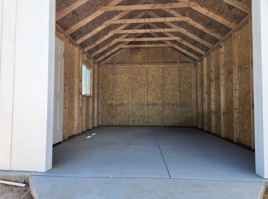 Interior view of an unfinished storage shed with wooden walls, an exposed truss ceiling, and a grey concrete floor.