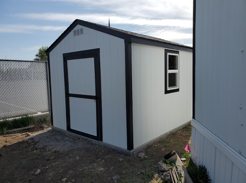 A white shed with black trim and a single window stands on a gravel base next to a chain-link fence.