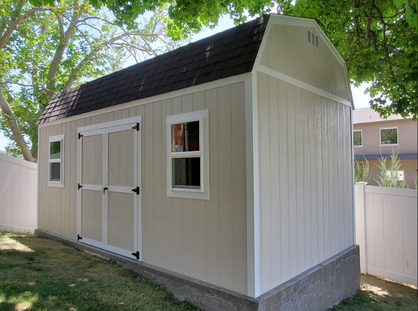 A beige wooden storage shed with a dark brown shingled barn-style roof, set in a grassy backyard next to a white fence.