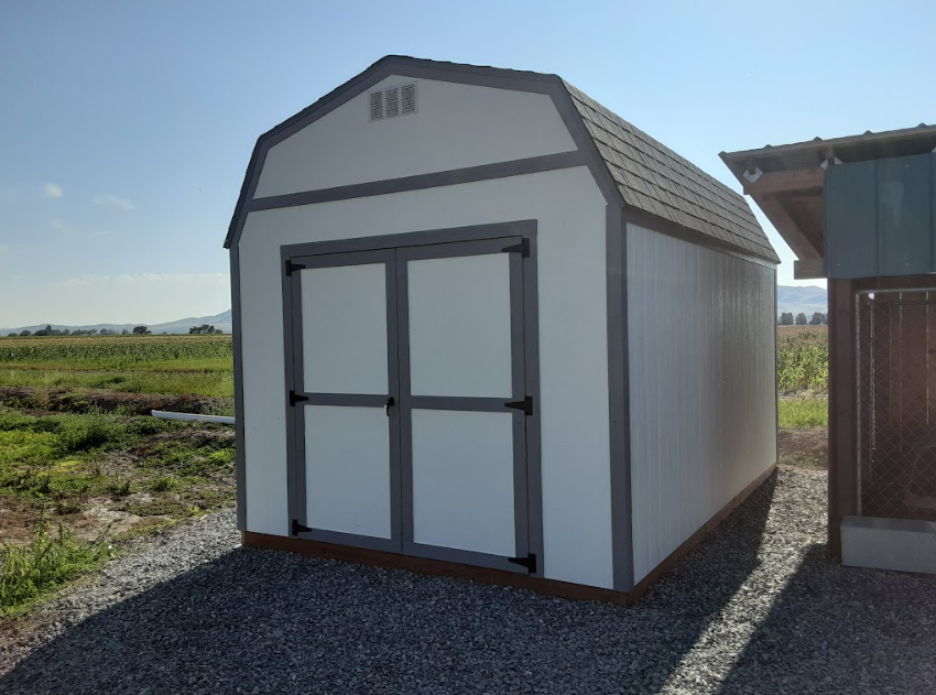 A white barn-style storage shed with gray trim sits on a gravel pad outdoors under a bright blue sky.