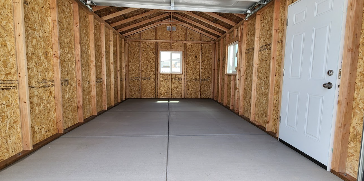 Interior of an unfinished wooden storage shed with grey flooring, a white door on the right, and two windows.