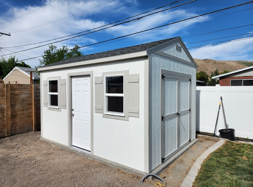 A white backyard shed with grey trim, a door, and two shuttered windows, set in a gravel and grass yard.