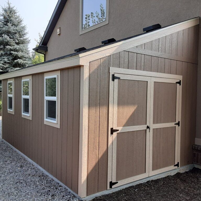 A brown shed with white trim and three windows is attached to the side of a larger house with a gravel ground in front.