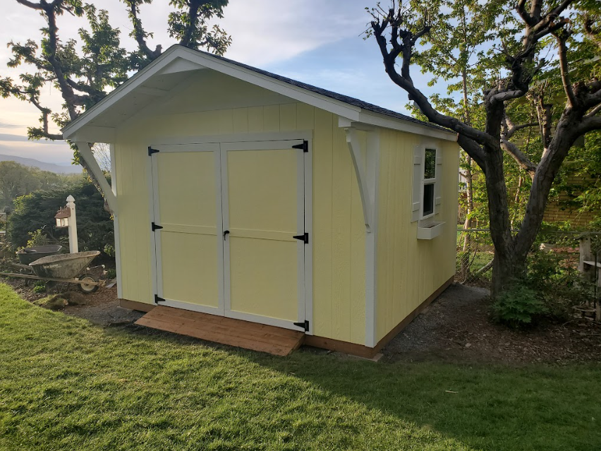 A light yellow shed with white trim and double doors sits on a grassy hill next to a large tree.