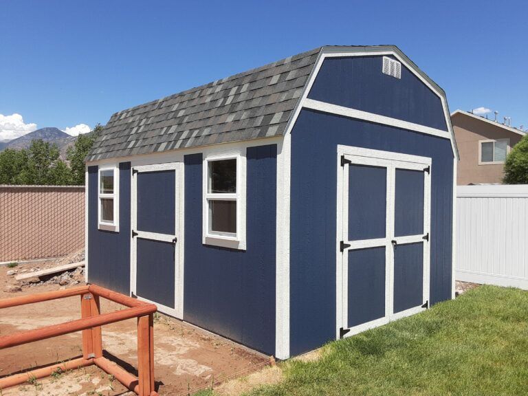 A navy blue gambrel-style shed with white trim, two windows, and double doors, situated in a backyard.