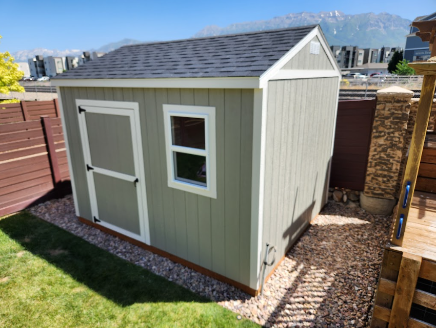 A sage-green storage shed with a white-trimmed door and window, set on a gravel base in a suburban backyard.