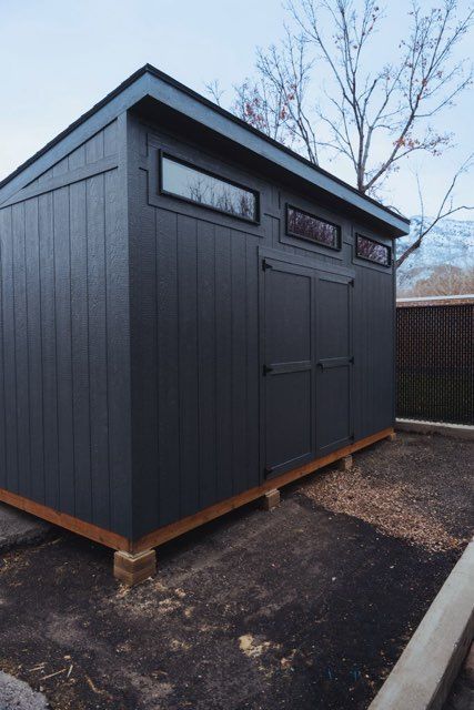 A modern, charcoal-gray shed with long horizontal windows and double doors, elevated on concrete blocks in a backyard.