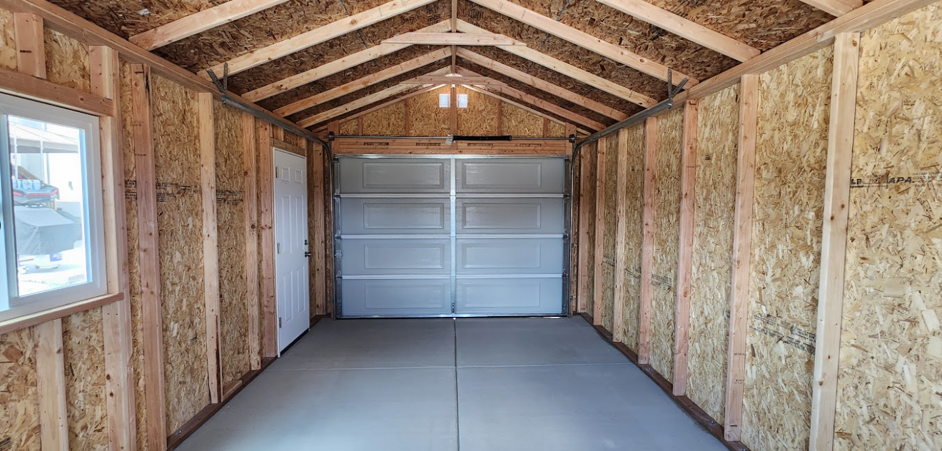 Interior view of an empty shed with bare wooden walls, a concrete floor, a side window, a door, and a garage-style door.
