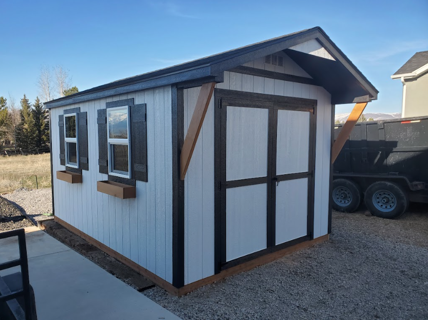 A white storage shed with dark brown trim, two windows with flower boxes, and double doors on a gravel lot.