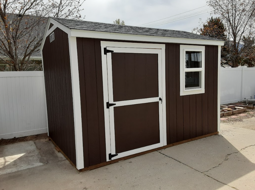 A brown wooden storage shed with a white trim, a single door, and one window, located on a concrete patio in a yard.