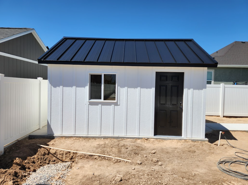 A white shed with a black door and black metal roof stands in a dirt yard between two white vinyl fences under a blue sky.