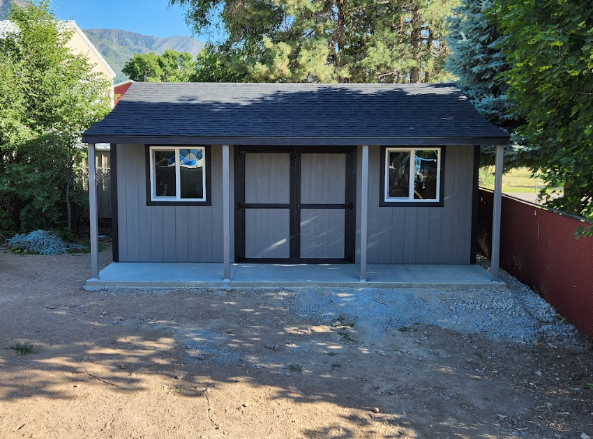 A gray wooden shed with black trim, a black shingled roof, two windows, and a front porch, situated on a dirt lot.