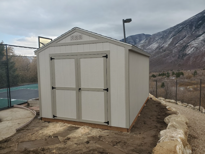 A beige wooden storage shed with double doors sits on a dirt plot next to a fence and a snowy mountain landscape.