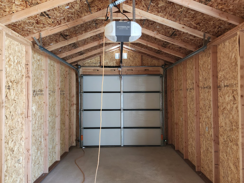 The interior of a wooden shed with an installed garage door opener hanging above a closed white roll-up door.