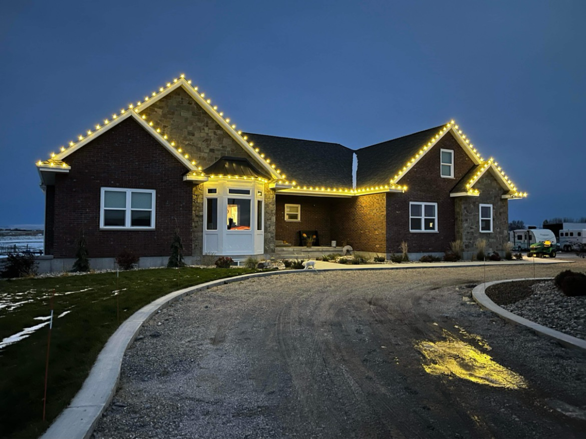 House with Christmas lights, yellow lights along roofline, brick exterior, dark sky.