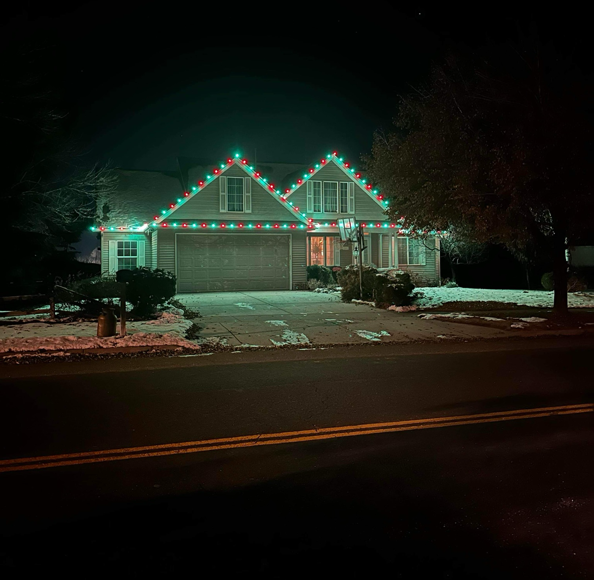 House at night with green and red Christmas lights on the roof, slight snow on ground.