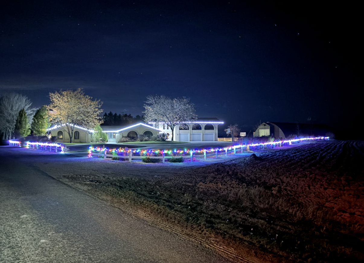 A house and fence are decorated with Christmas lights at night, in front of a dark field.