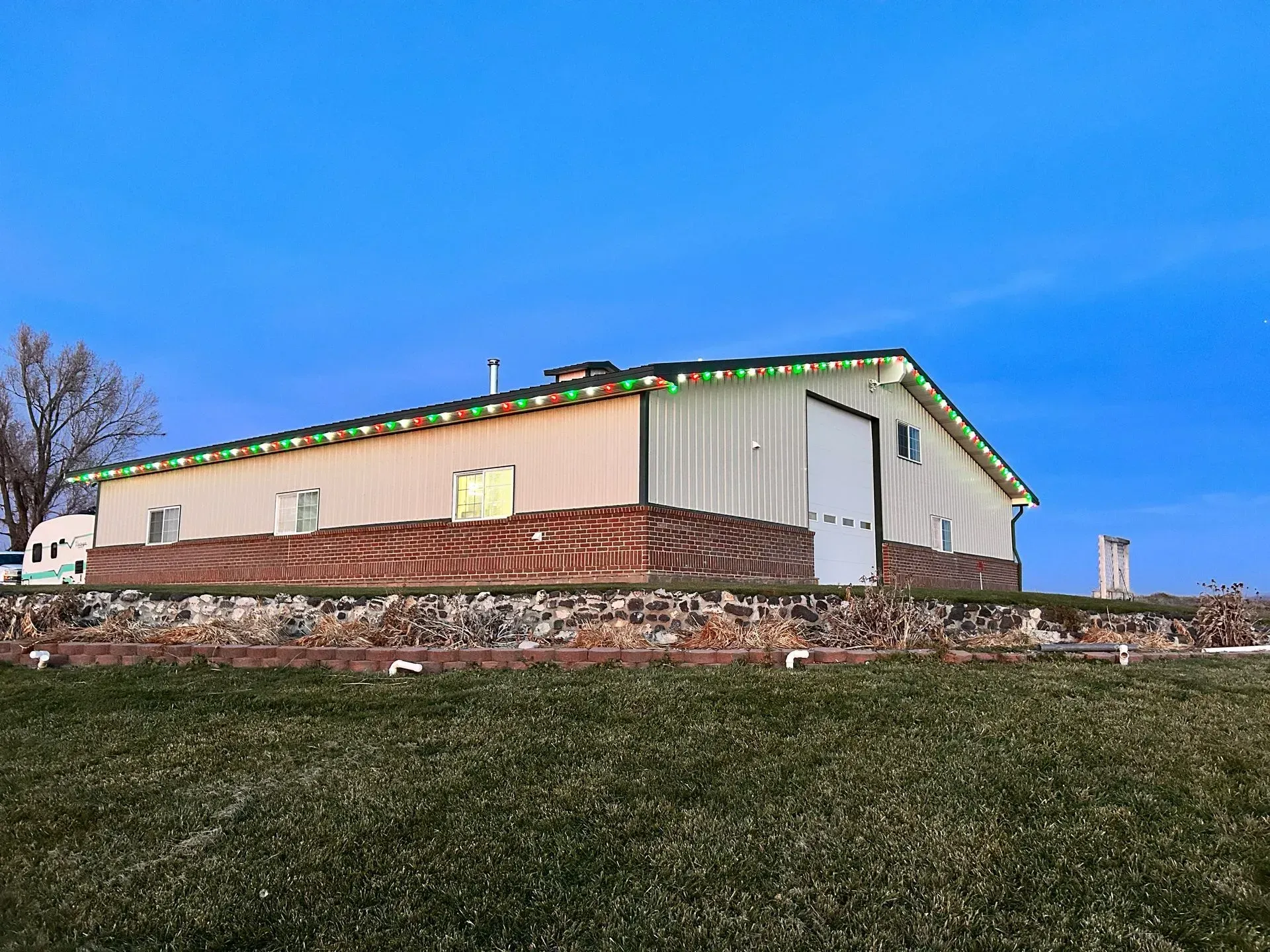 A large barn with christmas lights on the roof is sitting in the middle of a grassy field.