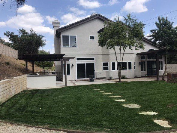 Backyard of a two-story house with green lawn, stone path, patio, and pergola structures.